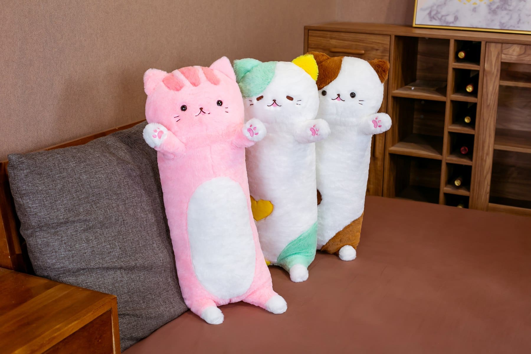 Auf einem grauen Sofa in einem kleinen Wohnzimmer mit braunem Boden liegen drei Plüschkissen mit einer großen Katze, die auf dem Sofa lehnen, eines ist rosa, das zweite grün und das dritte braun