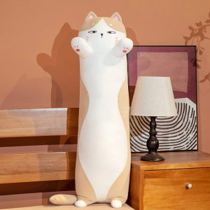Auf einem Bett, neben dem sich ein Nachttisch mit einer weißen Nachttischlampe und einem Rahmen dahinter befindet, liegt ein weißes und braunes Katzenkissen, das auf seinen winzigen Hinterpfoten steht