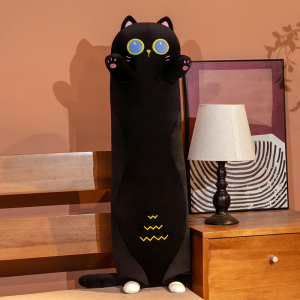 Auf einem Bett, neben dem ein Nachttisch mit einer weißen Nachttischlampe und einem Rahmen dahinter steht, befindet sich ein Plüschkissen einer schwarzen Katze, die auf ihren winzigen Hinterbeinen steht